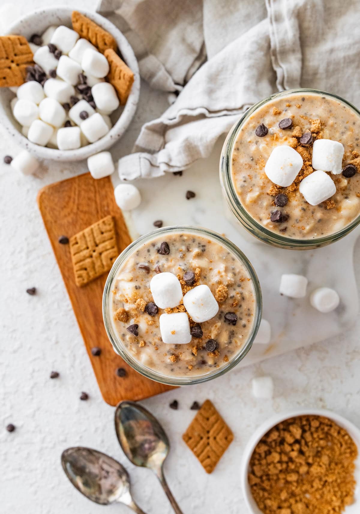 Two glass jars of s'mores overnight oats near smaller bowls with crushed graham cracker and mini marshmallows.