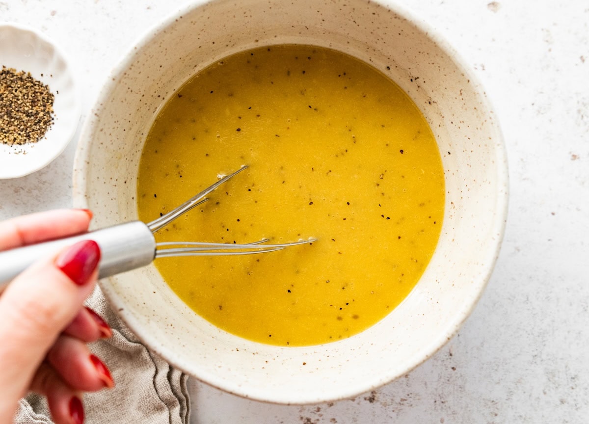 A womans hand uses a metal whisk to whisk an apple cider vinegar dressing in a bowl.