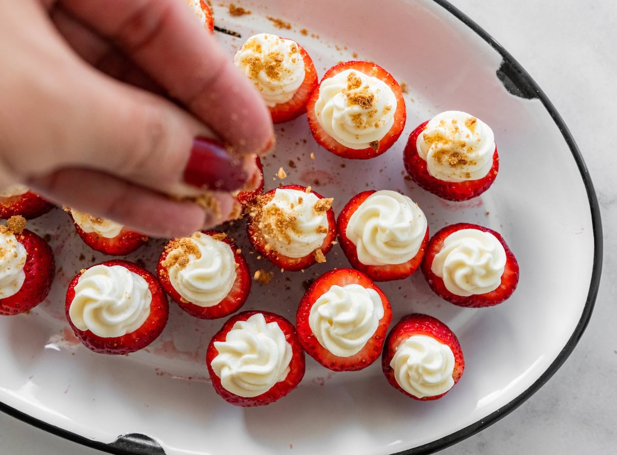 A womans hand sprinkles crushed graham cracker over strawberries stuffed with a cream cheese and yogurt mixture.