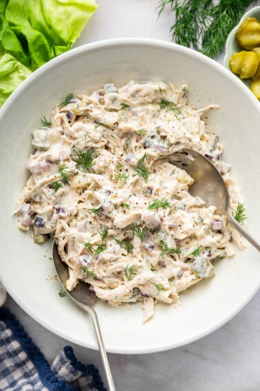 Dill pickle chicken salad in a large serving bowl with two serving spoons and topped with fresh dill.