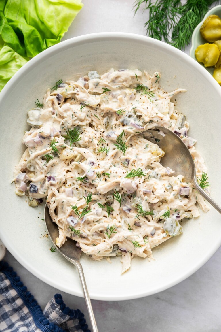 Dill pickle chicken salad in a large serving bowl with two serving spoons and topped with fresh dill.