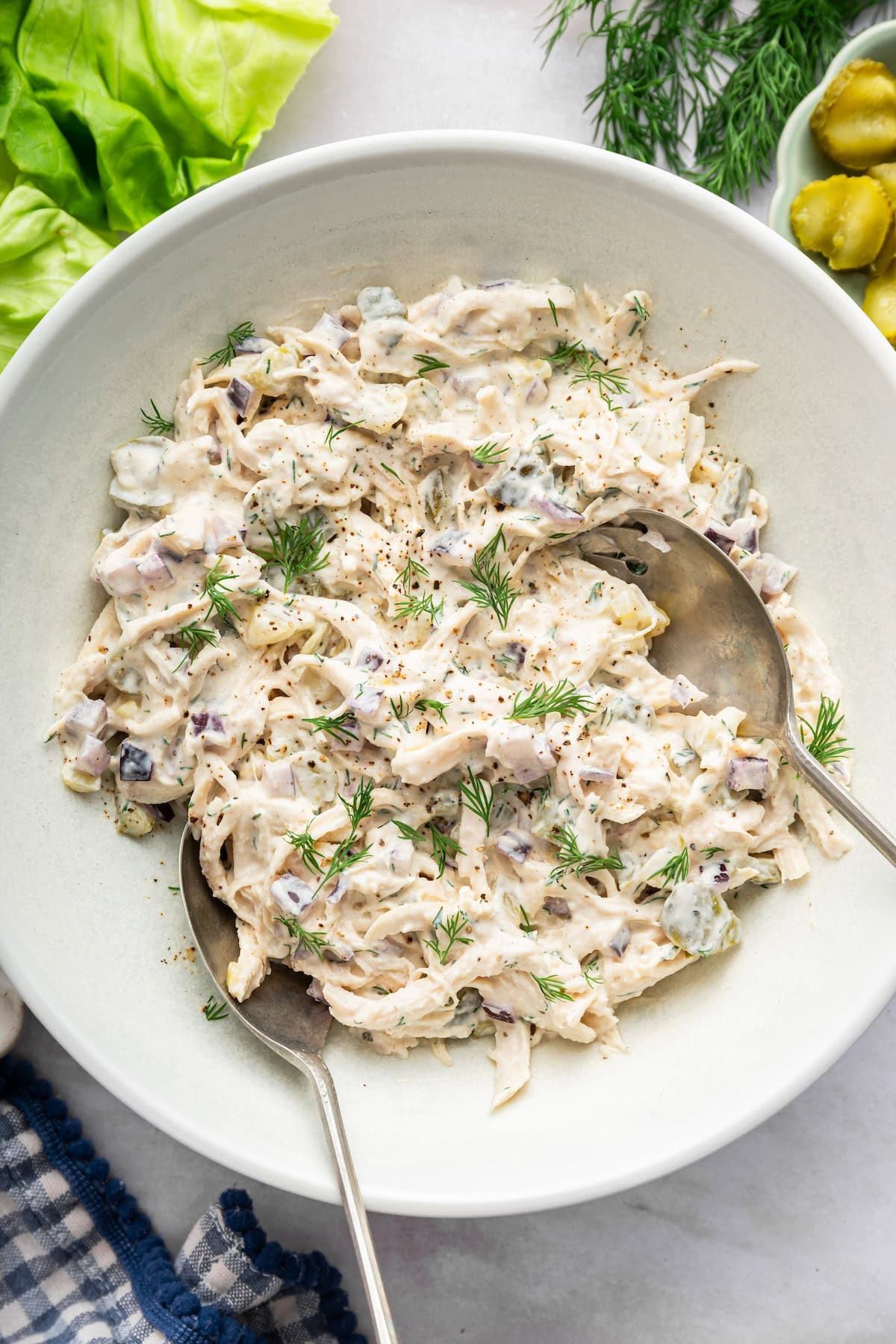 Dill pickle chicken salad in a large serving bowl with two serving spoons and topped with fresh dill.