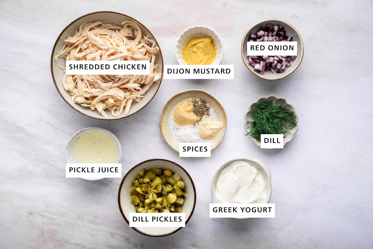 Ingredients measured out for dill pickle chicken salad: shredded chicken, dijon mustard, red onion, dill, greek yogurt, dill pickles, pickle juice, and spices.