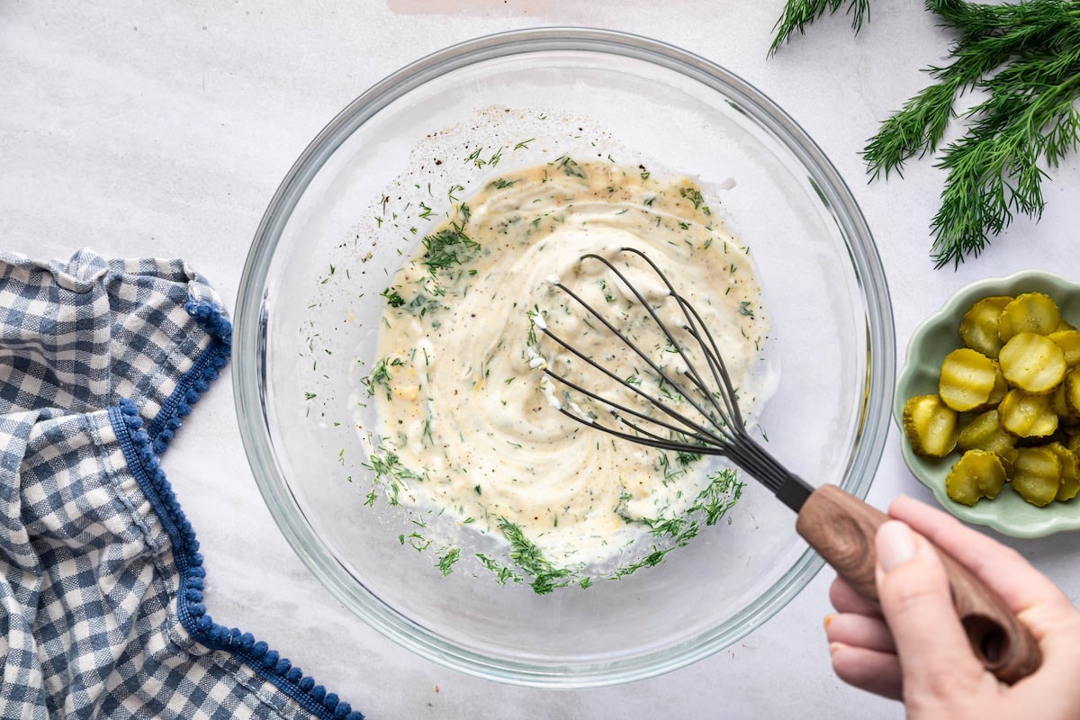A hand uses a whisk to mix a Greek yogurt and fresh dill dressing.