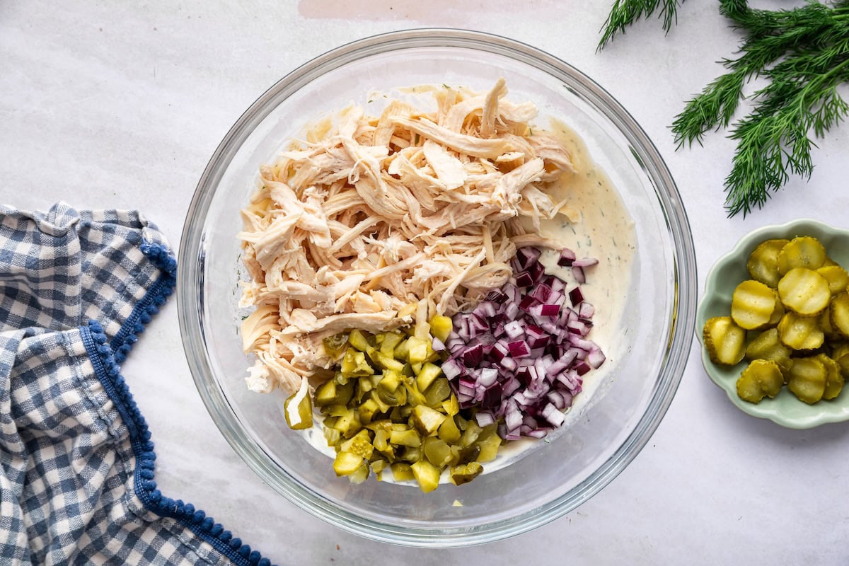Shredded chicken, dill pickles, and diced red onion in a large glass bowl with a creamy Greek yogurt sauce underneath.