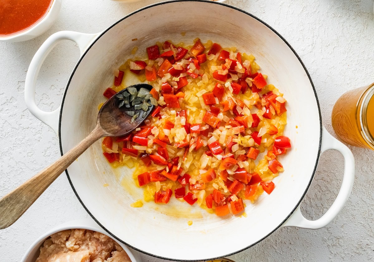 Yellow onion, garlic and red bell pepper sautéing in a dutch oven with a wooden spoon.