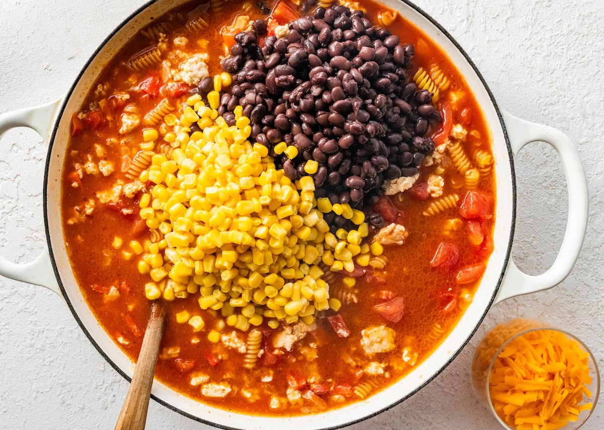 Corn and black beans being added to the Dutch oven with the rest of the enchilada pasta ingredients.