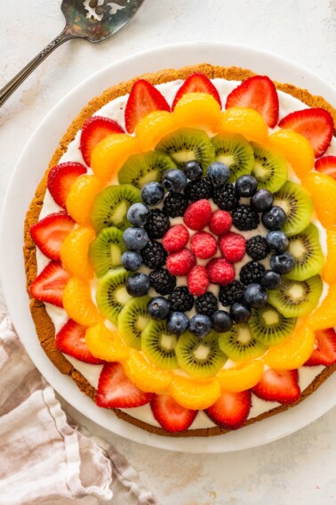 Sugar cookie fruit pizza topped with strawberries, mandarin oranges, kiwi, blueberries, blackberries and raspberries arranged in a colorful pattern.