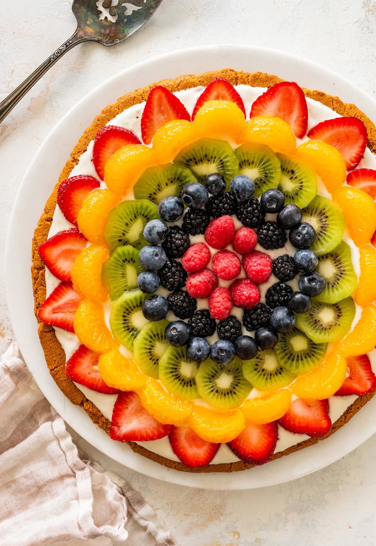 Sugar cookie fruit pizza topped with strawberries, mandarin oranges, kiwi, blueberries, blackberries and raspberries arranged in a colorful pattern.
