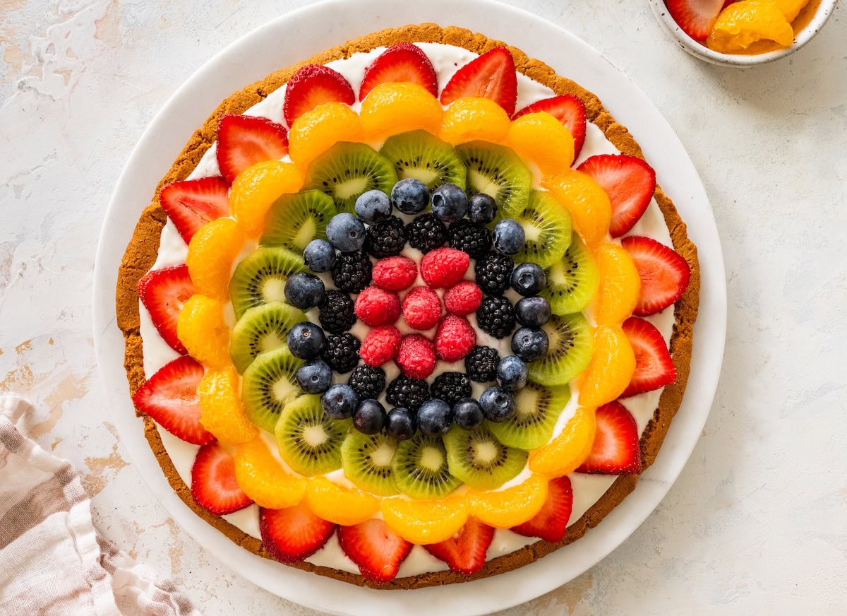 Fruit pizza with vibrant layers of fresh fruit arranged in a circular design (strawberries, mandarin oranges, kiwi, blueberries, blackberries and raspberries).