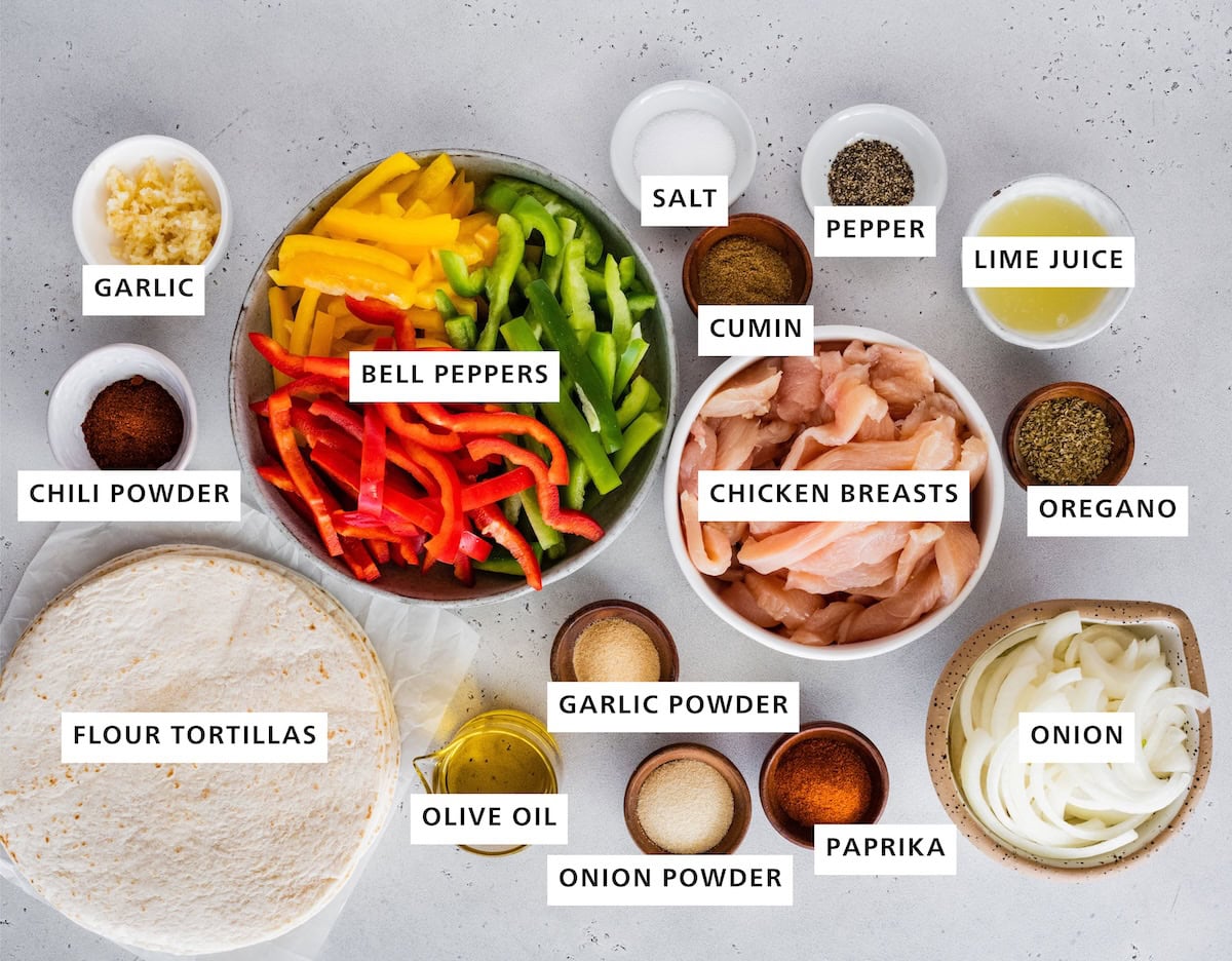 Ingredients measured out to make Healthy Chicken Fajitas: flour tortillas, chili powder, garlic, tri-colored peppers, olive oil, garlic powder, onion powder, paprika, chicken breasts, cumin, sea salt, pepper, lime juice, oregano and yellow onion.