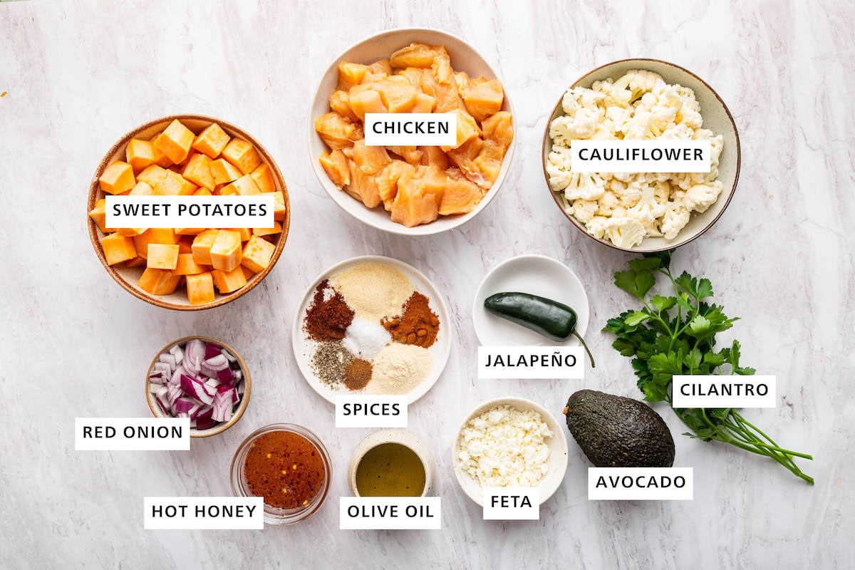 Ingredients measured in small dishes for hot honey chicken sheet pan meal: chicken, cauliflower, sweet potatoes, red onion, spices, hot honey, olive oil, feta, jalapeño, avocado, and cilantro.