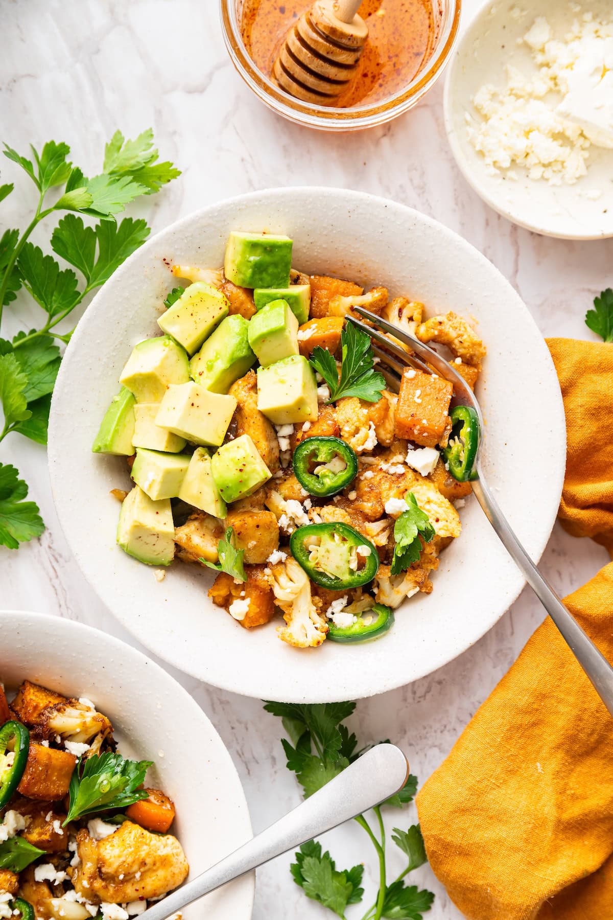 A serving of hot honey chicken sheet pan meal in a bowl topped with avocado chunks, sliced jalapeño, fresh herbs, and feta cheese.