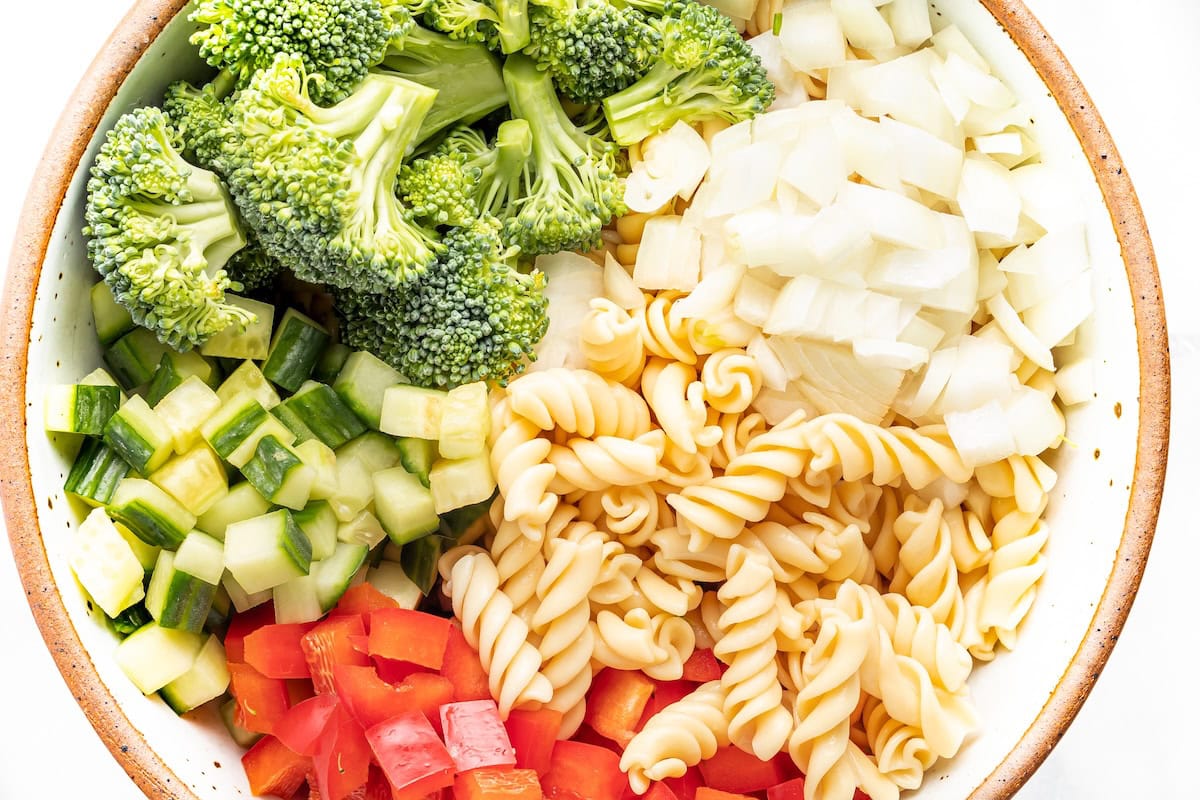 Red bell pepper, cucumber, broccoli, onion and pasta in a bowl before being tossed together.