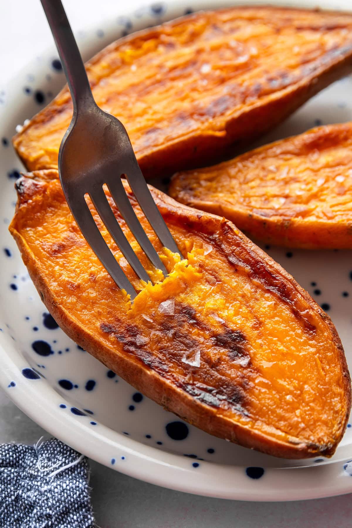 Three oven roasted sweet potato halves on a plate sprinkled with flaky sea salt. There is a fork piercing one of the halves.