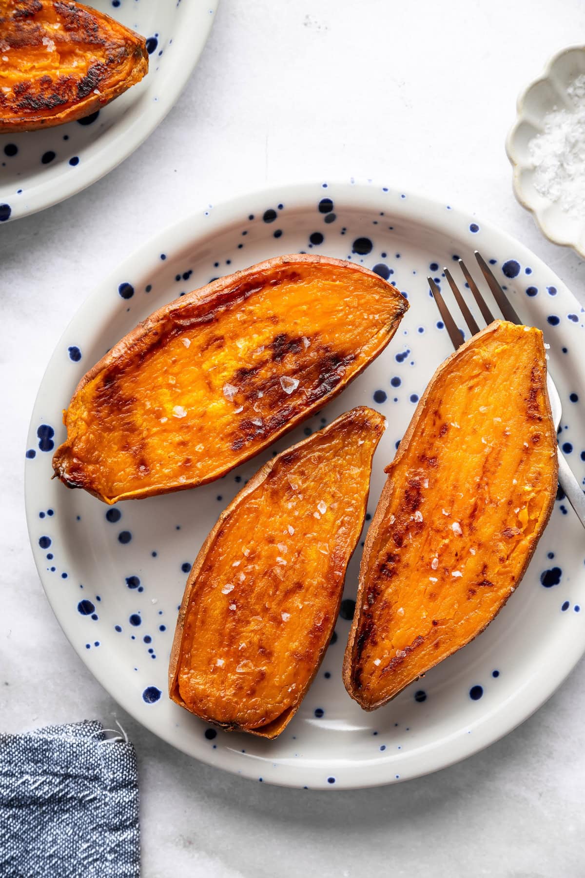 Three oven roasted sweet potato halves on a blue and white speckled plate sprinkled with flaky sea salt.