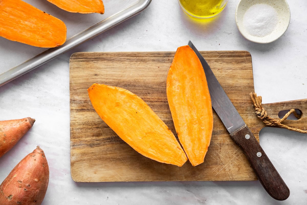 A sweet potato cut in half on a wooden cutting board next to a knife.