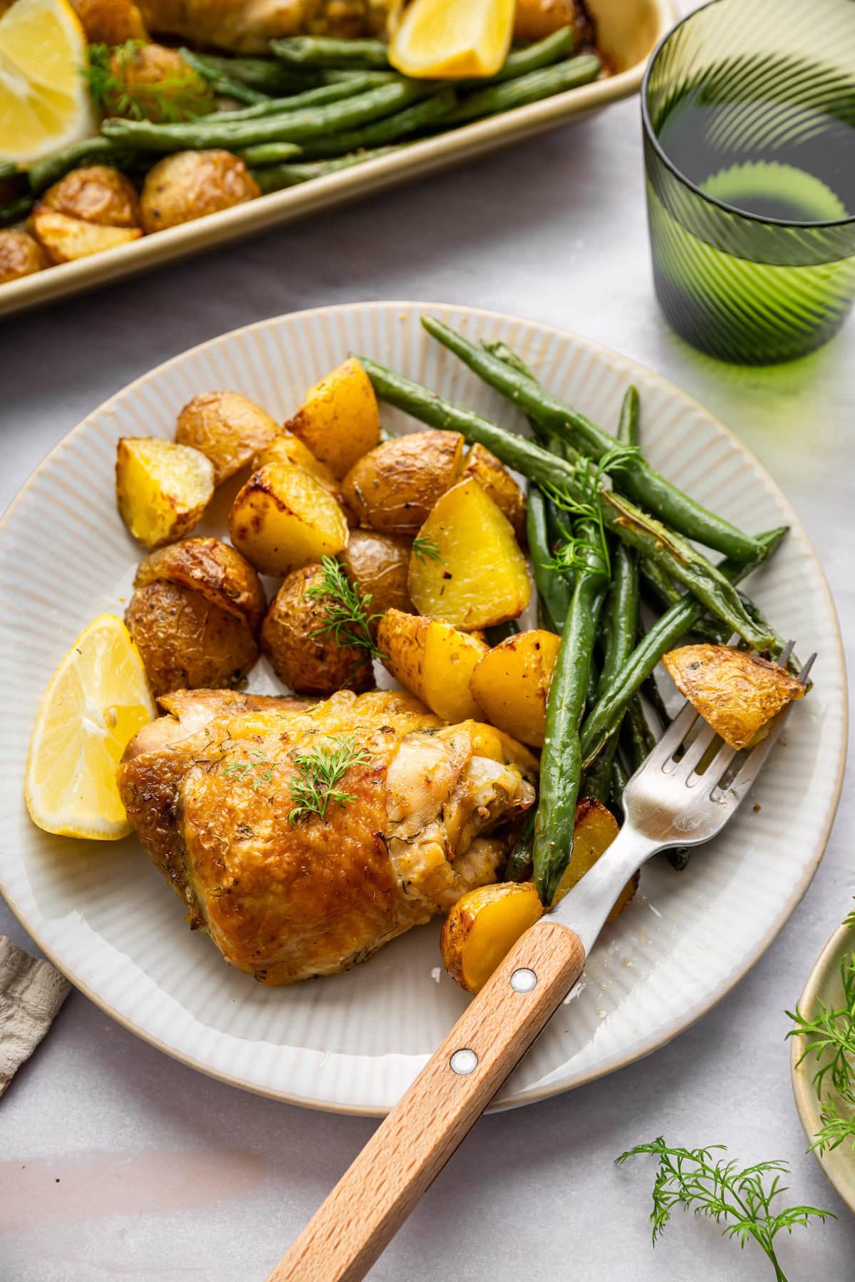 A plate with a baked chicken thigh, roasted potatoes, green beans, and a lemon wedge on the side.