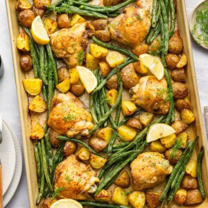 A sheet pan with baked chicken thighs, green beans, potatoes, fresh dill, and lemon wedges.