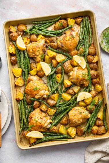 A sheet pan with baked chicken thighs, green beans, potatoes, fresh dill, and lemon wedges.