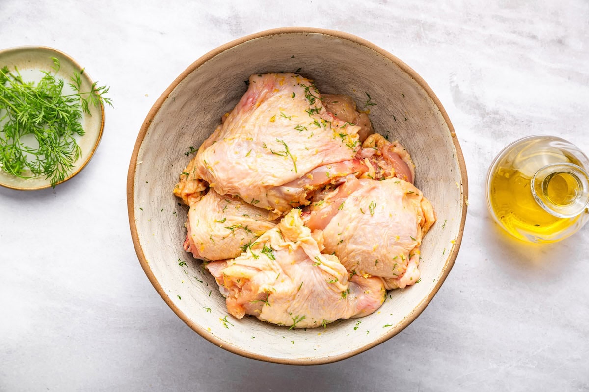 Raw chicken thighs in a large bowl with olive oil and fresh dill.