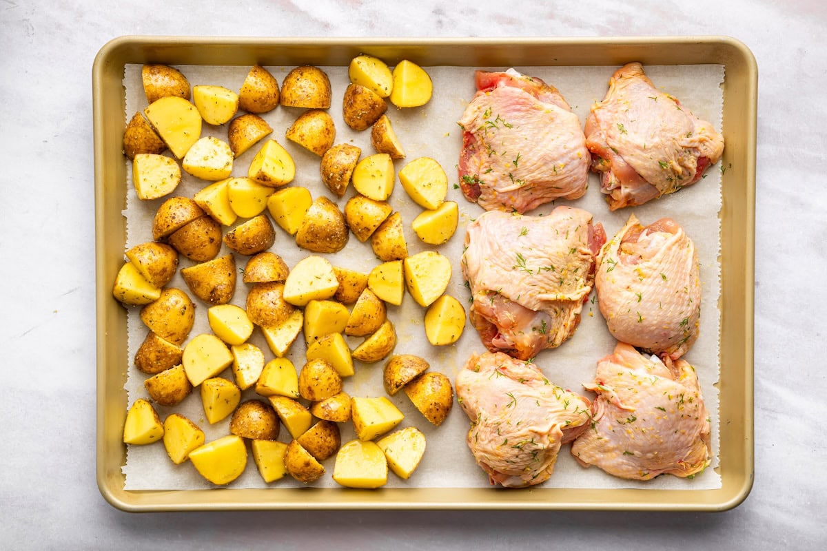 Raw chicken thighs on one side of a sheet pan and potatoes on the other side before going in the oven.
