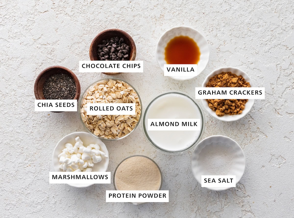 Ingredients measured out for s'mores overnight oats: rolled oats, almond milk, graham crackers, sea salt, protein powder, marshmallows, chia seeds, chocolate chips, and vanilla.
