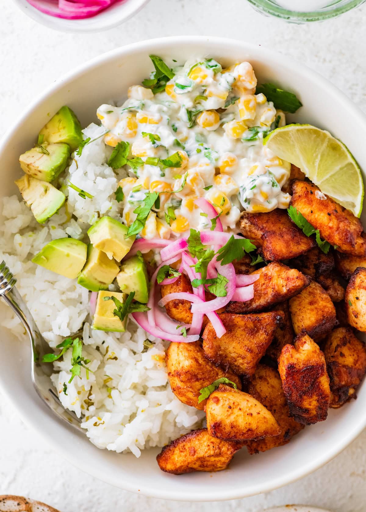 Seasoned chicken, Mexican street corn, cilantro lime rice, avocado, pickled red onion, and a lime wedge in a bowl.