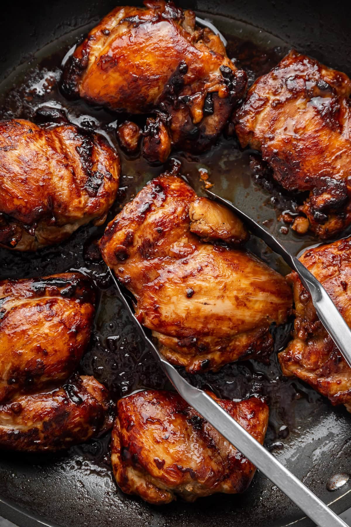 Metal tongs grabbing a chicken thigh from a large skillet with more teriyaki chicken thighs.
