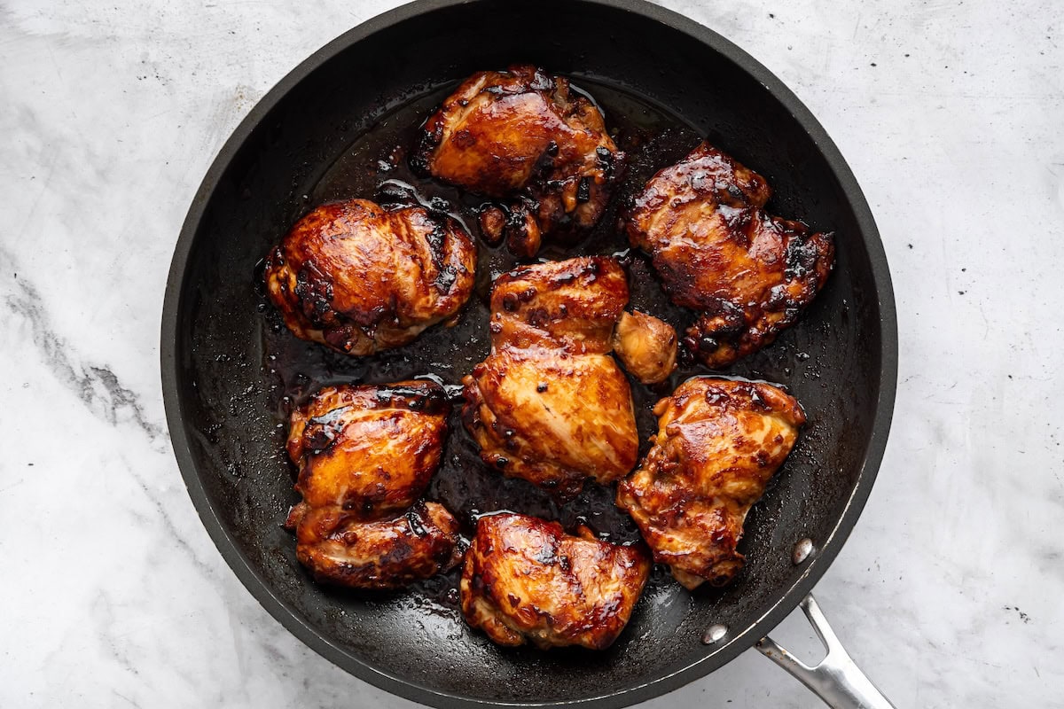 Six chicken thighs with a teriyaki marinade cooking in skillet.