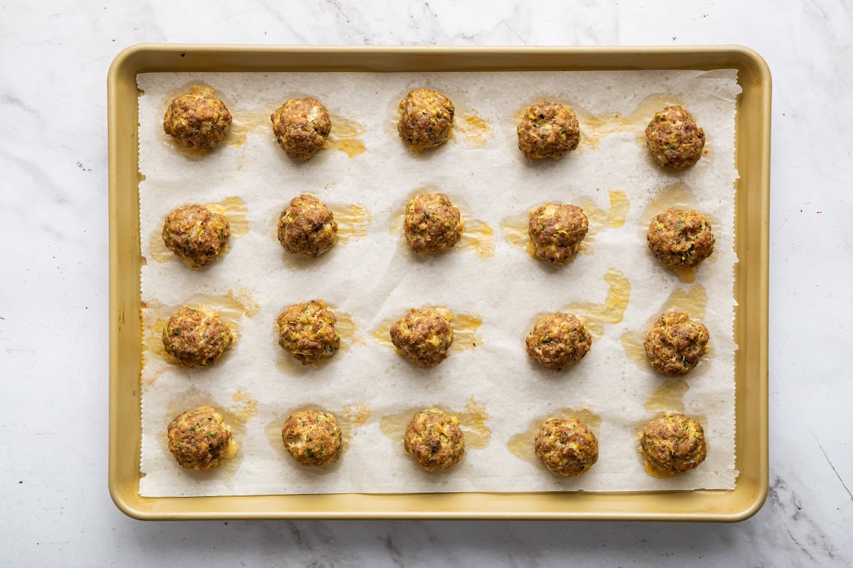 Zucchini meatballs spread out on a baking tray after being baked in the oven.
