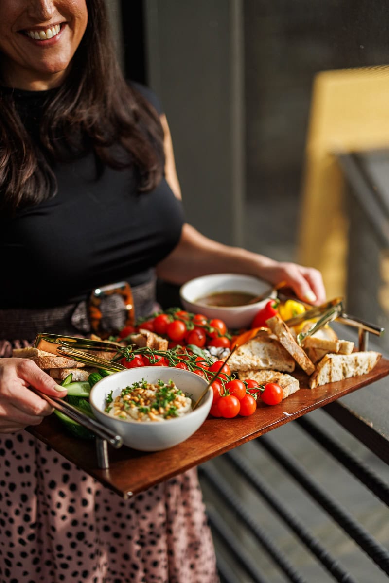 Woman carrying a Mediterranean appetizer tray with hummus, pita, tomatoes, cucumbers, and peppers.