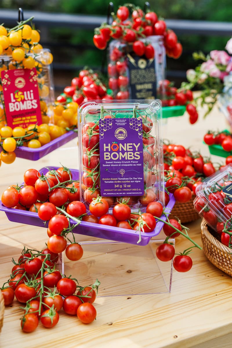 Display of packaged Honey Bombs cherry tomatoes surrounded by fresh tomatoes on the vine.