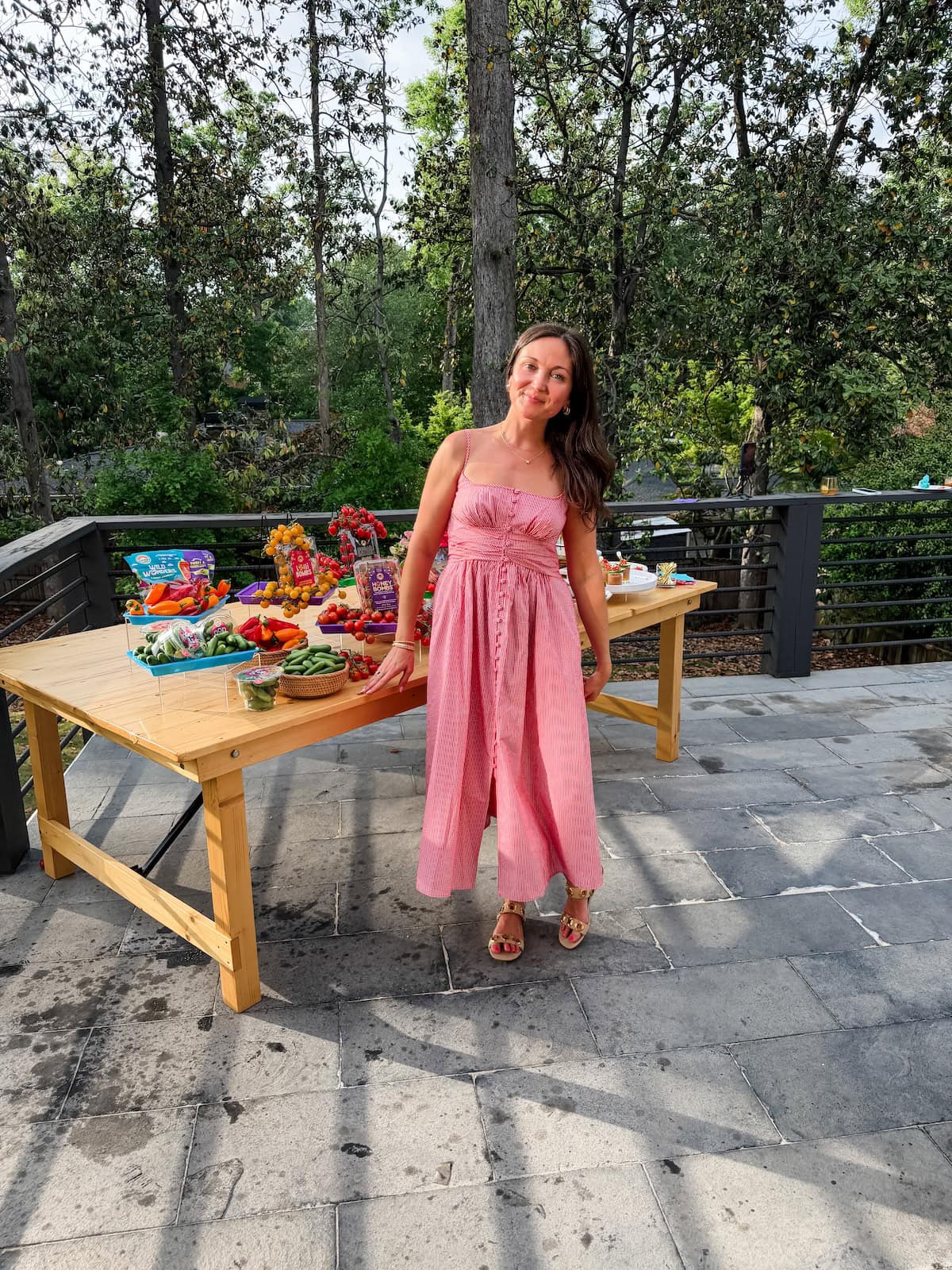Woman in a pink dress standing beside a fresh tomato and vegetable display on an outdoor patio.