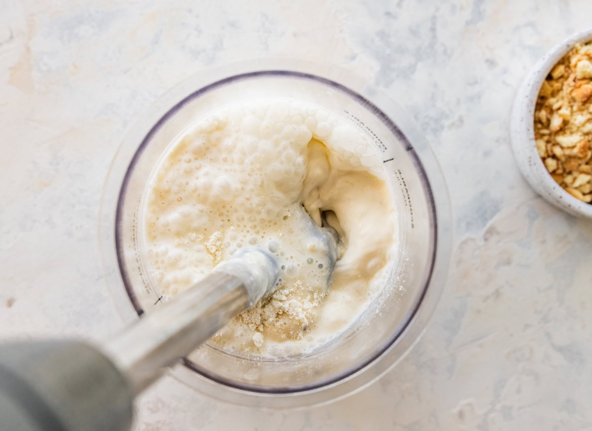 An immersion blender in a container blending banana and protein powder.