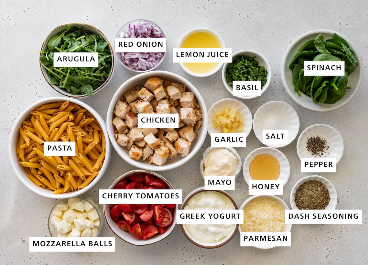 Ingredients measured out for chicken pesto pasta salad: arugula, red onion, lemon juice, basil, spinach, pepper, dash seasoning, parmesan, honey, salt, garlic, mayo, greek yogurt, cherry tomatoes, chicken, pasta, and mozzarella balls.
