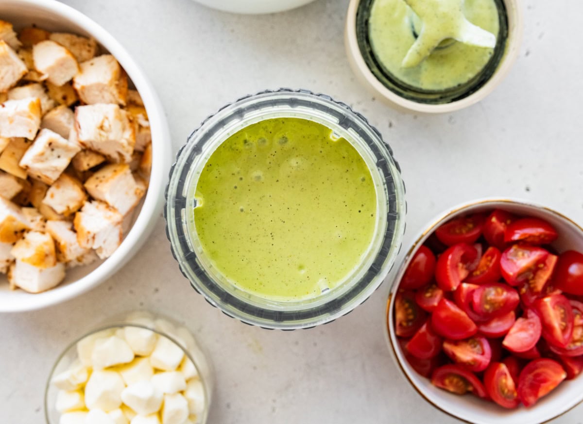 A pesto dressing blended in a small blender with bowls of cooked chopped chicken and cherry tomatoes nearby.
