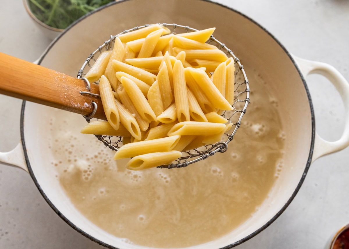Cooked pasta held over a pot of boiling water by a strainer spoon.