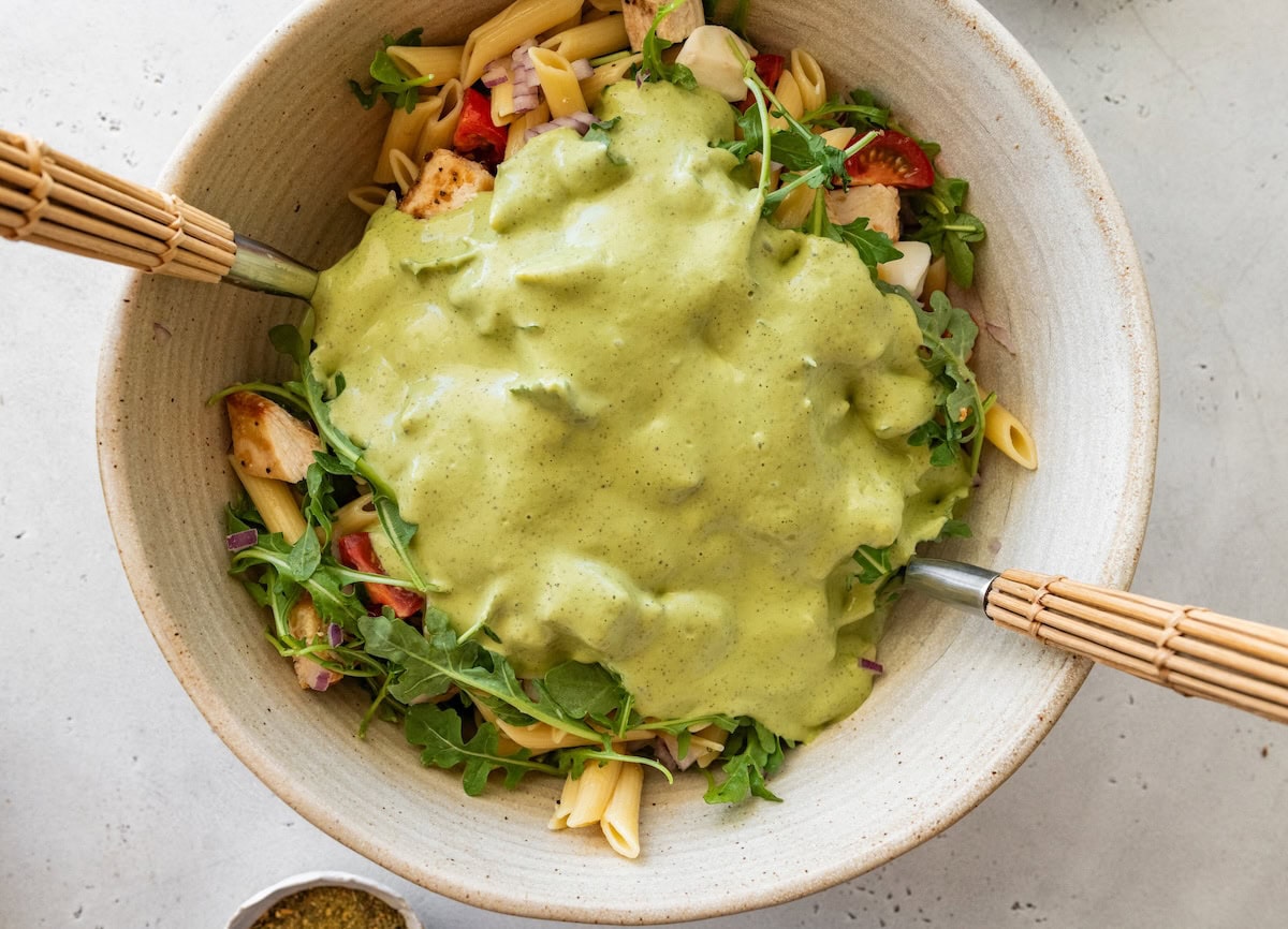 Pesto dressing added to a large mixing bowl of chicken pesto pasta salad with two serving spoons in the bowl.