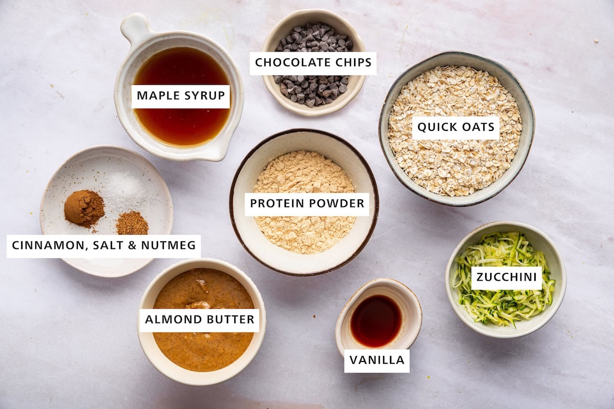 Ingredients measured out for zucchini bread protein balls: protein powder, quick oats, zucchini, vanilla, almond butter, cinnamon, salt, nutmeg, maple syrup, and chocolate chips.