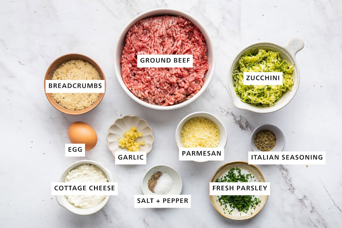 Ingredients measured out for zucchini meatballs: ground beef, zucchini, Italian seasoning, fresh parsley, salt, pepper, cottage cheese, egg, breadcrumbs, garlic, and parmesan.