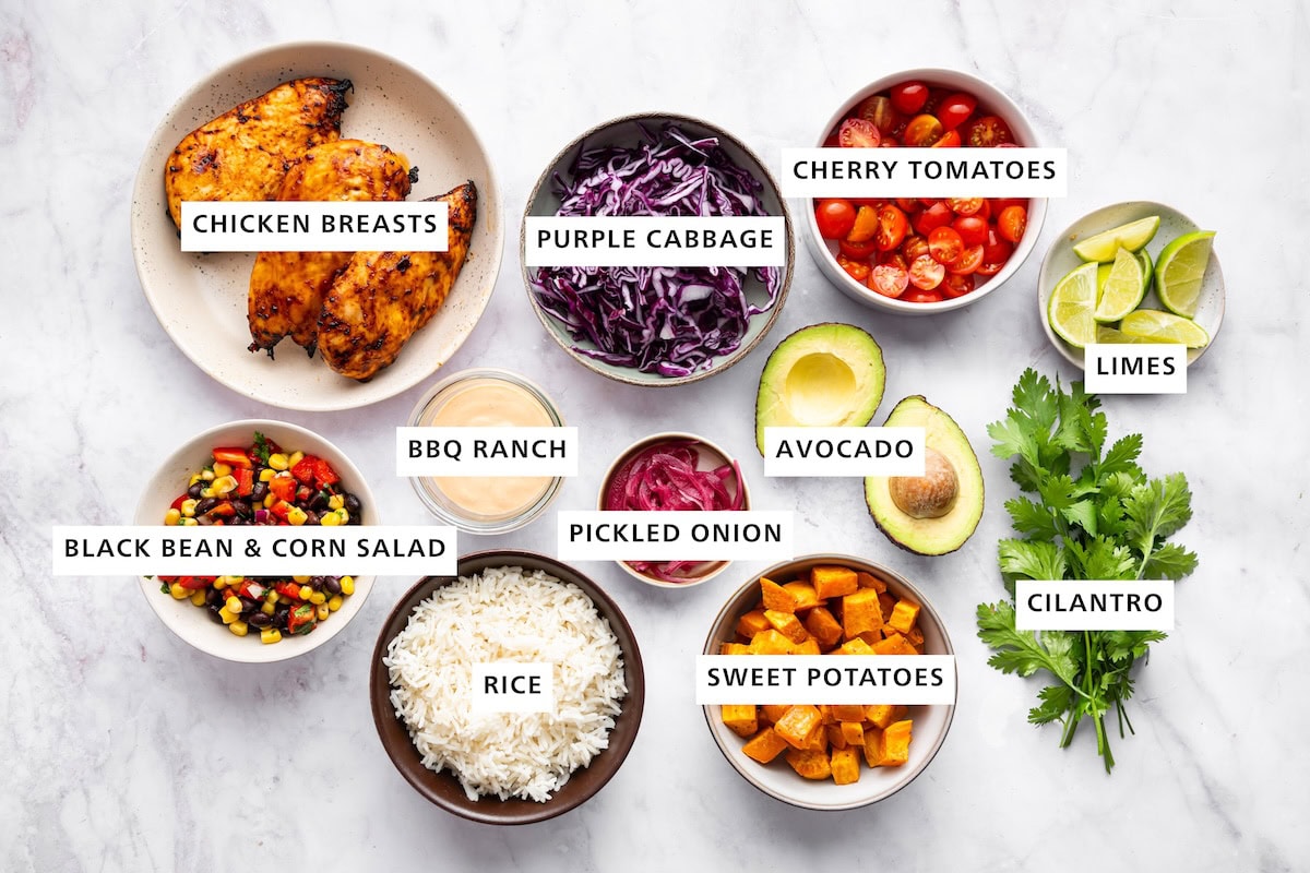 Ingredients measured out for BBQ chicken bowls: chicken breasts, purple cabbage, cherry tomatoes, limes, cilantro, pickled onion, avocado, sweet potatoes, rice, black bean corn salad, and BBQ ranch