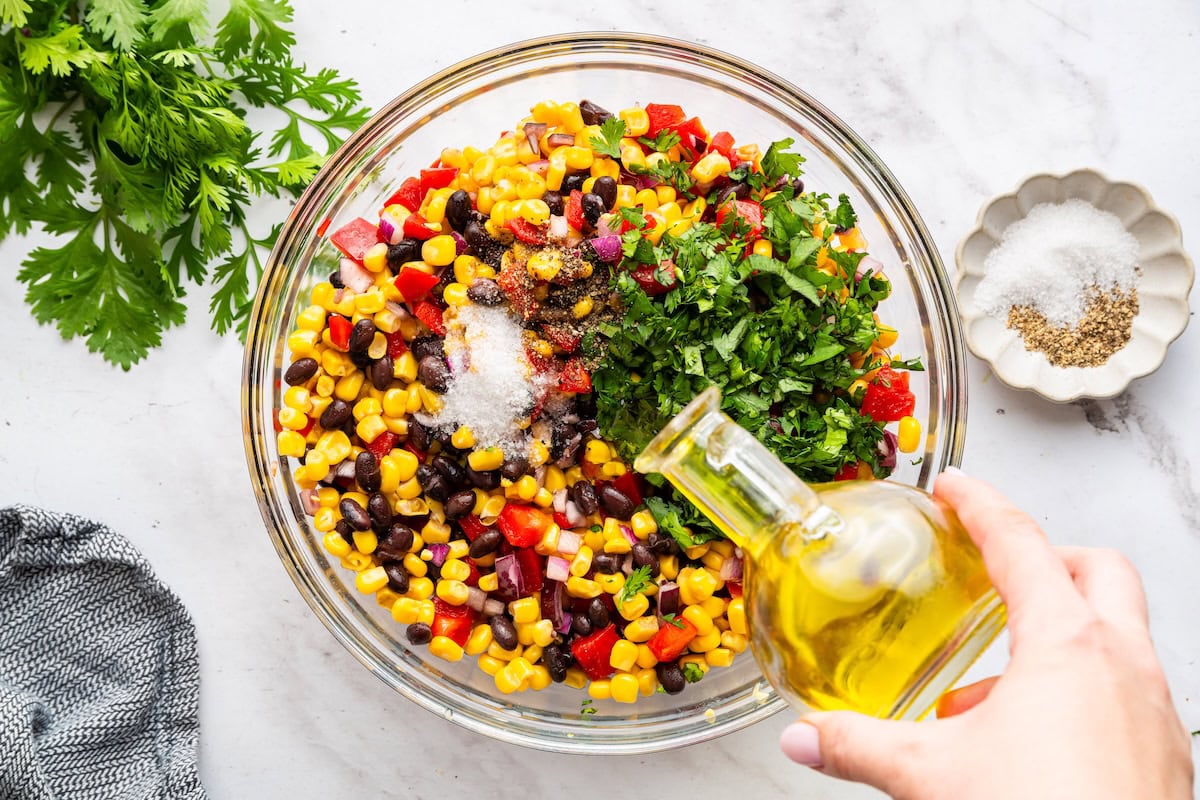 A woman's hand pours a glass container of oil in a large glass bowl of black bean and corn salad.