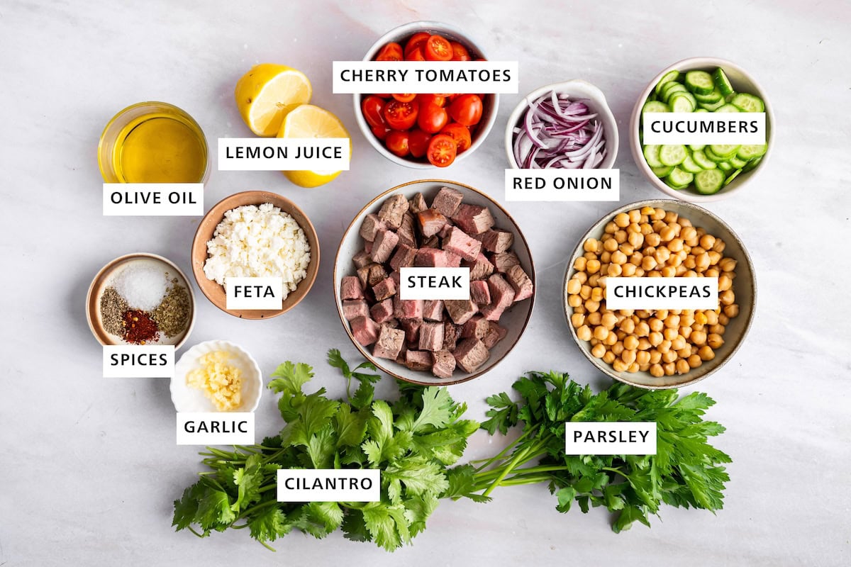 Ingredients measured out for chimichurri steak salad including: steak, chickpeas, parsley, cilantro, garlic, spices, feta, olive oil, lemon juice, cherry tomatoes, red onion, cucumbers.