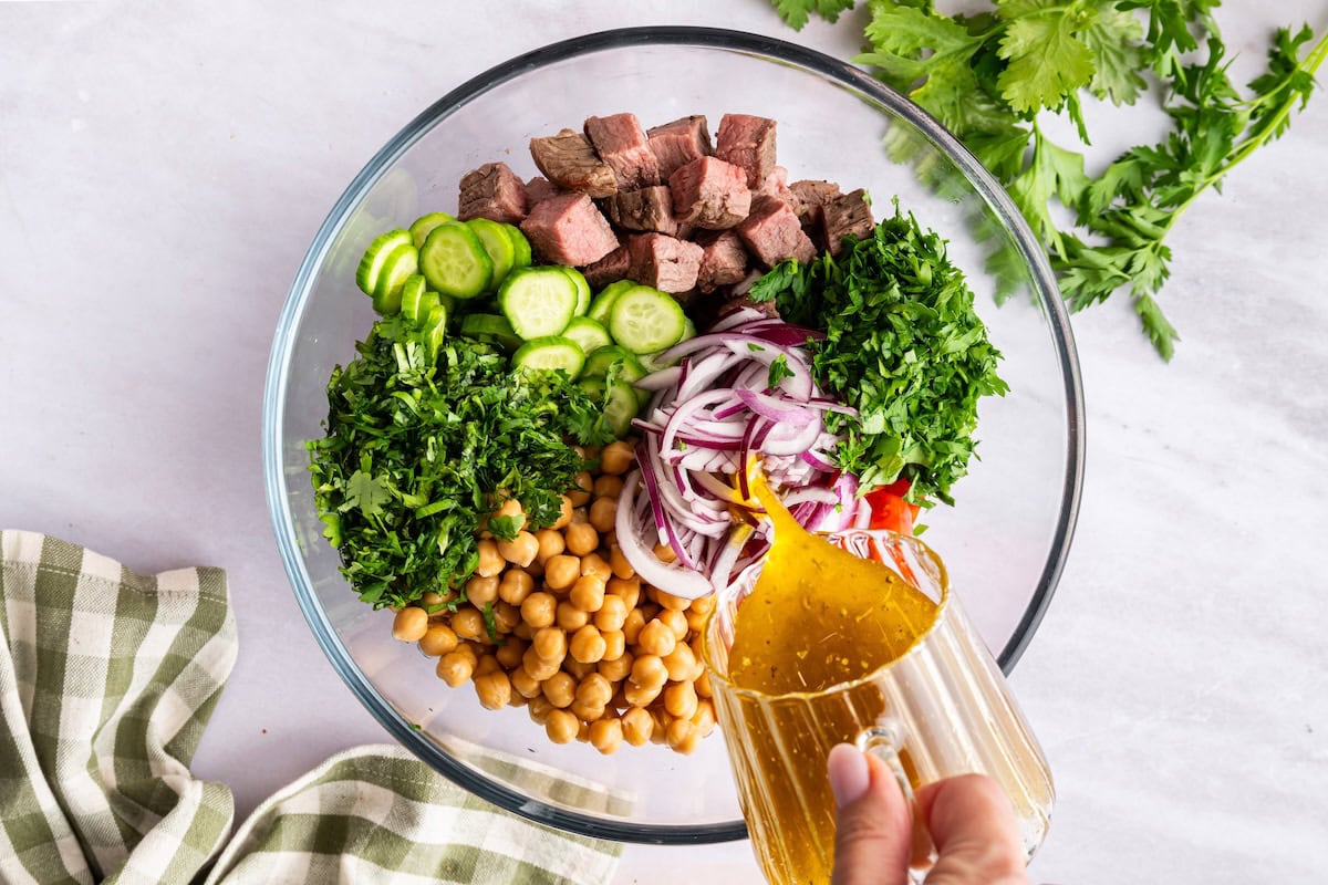 Dressing is poured into a large glass bowl of ingredients used for chimichurri steak salad.