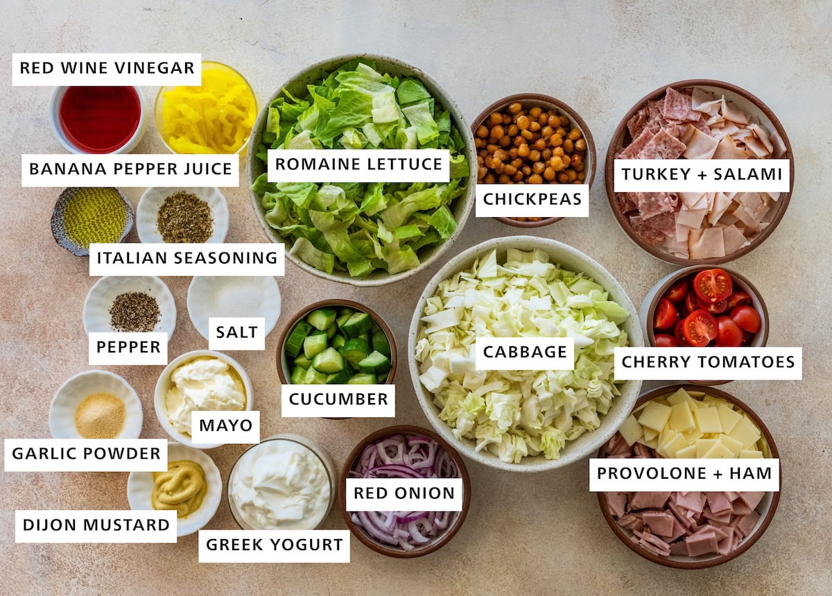 Ingredients measured out for a grinder salad including: romaine lettuce, chickpeas, turkey, salami, cherry tomatoes, provolone, ham, cabbage, red onion, cucumber, greek yogurt, mayo, dijon mustard, garlic powder, pepper, salt, italian seasoning, banana pepper juice, red wine vinegar.