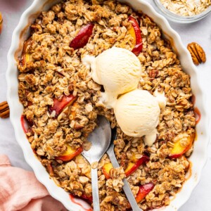 Peach crisp in a large oval baking dish served with two scoops of vanilla ice cream.