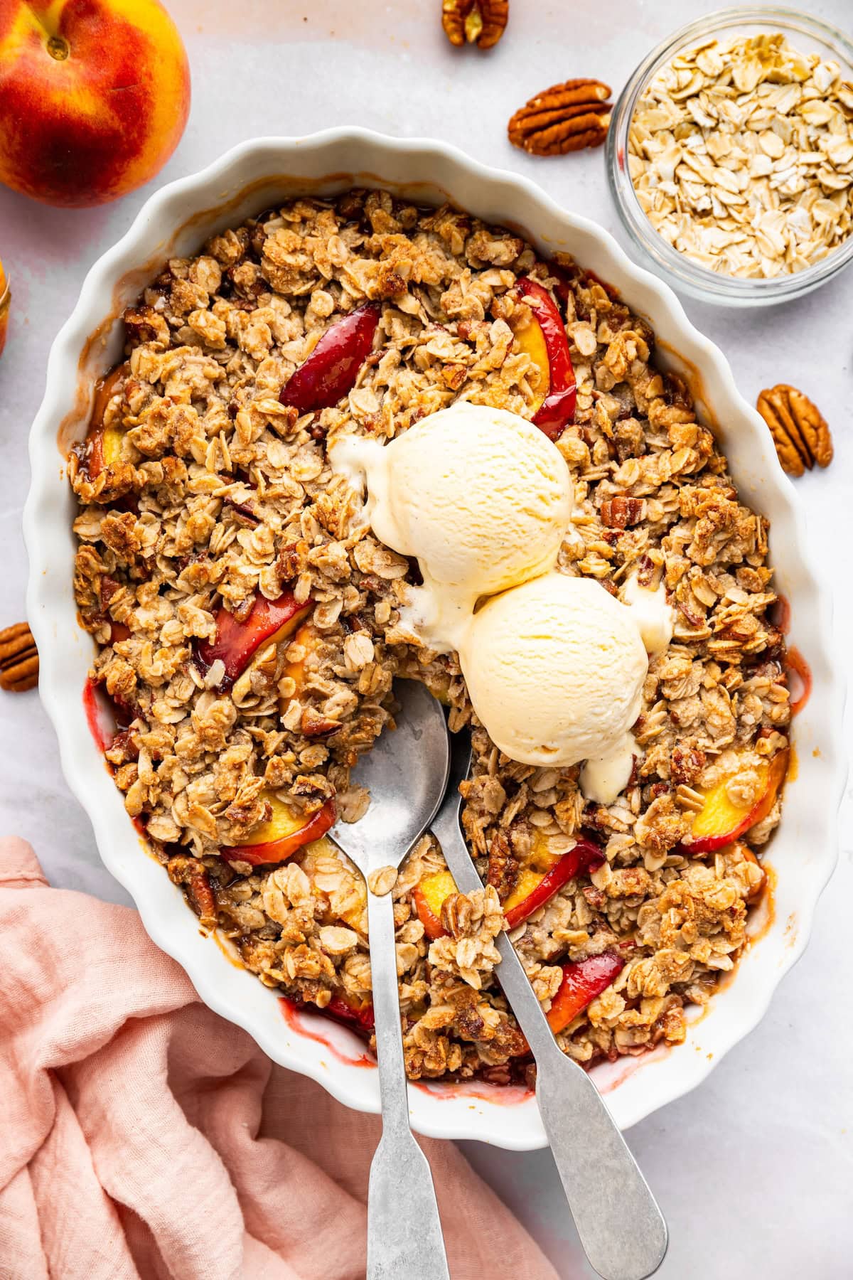 Peach crisp in a large oval baking dish served with two scoops of vanilla ice cream.