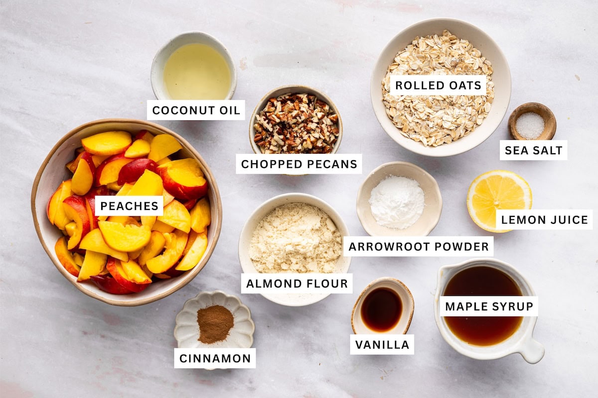 Ingredients measured out for a healthy peach crisp: peaches, cinnamon, almond flour, vanilla, maple syrup, lemon juice, sea salt, rolled oats, arrowroot powder, chopped pecans, and coconut oil.