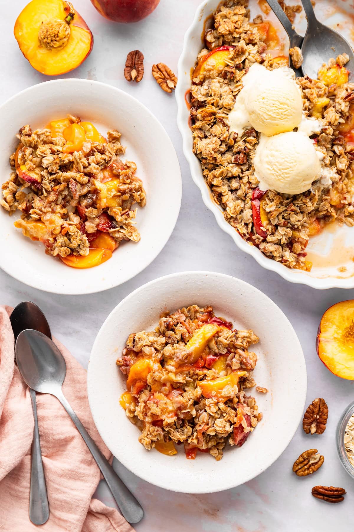 Two servings of peach crisp in bowls near a large baking dish of the peach crisp.